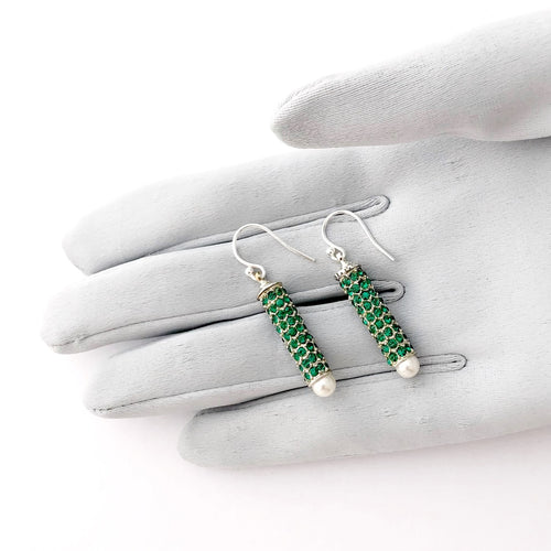 cylindrical earrings set with green rhinestones and a white pearl bead displayed on a gloved hand