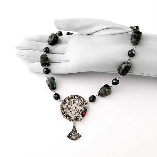 necklace in silvery greys with a red rhinestone set focal displayed on a gloved hand