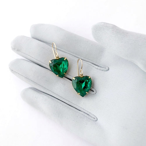 a pair of large emerald green heart shaped rhinestone earrings on a gloved hand